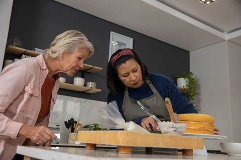 Mature women collaborating while decorating layered sponge cake in modern kitchen
