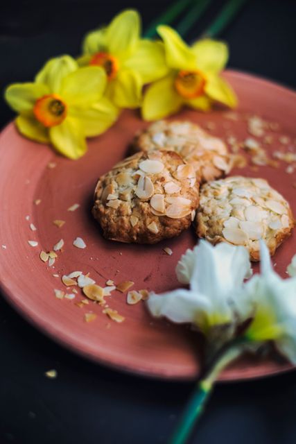 Oatmeal almond cookies placed on a pink plate with yellow daffodil flowers nearby. This vibrant and aesthetically pleasing arrangement is perfect for use in food blogs, culinary websites, spring-themed content, baking advertisements, and lifestyle magazines. The image captures the essence of homemade baking with a seasonal, rustic touch.