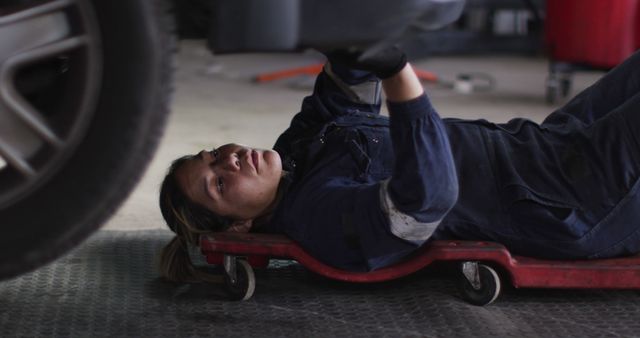 Depicts female mechanic working underneath a car on a creeper in a garage setting. Ideal for promoting gender diversity in traditionally male-dominated professions, automotive service advertisements, or educational materials related to car repair and maintenance.