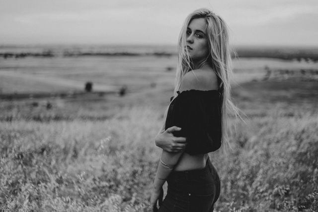 A young blonde woman with long hair is standing in an open field, looking directly at the camera. She is wearing a casual outfit, including an off-shoulder top and jeans. The photo is in black and white, giving it a timeless and classic feel. This image can be used to convey a sense of calm, introspection, nature, rural life, simplicity, or casual fashion.