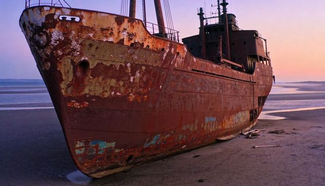Rusted cargo ship resting on tidal flat at sunset with peeling paint and driftwood debris