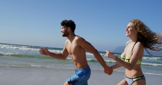 A young Caucasian man and woman are enjoying a run along the shore of a sunny beach, with copy space. Their joyful expressions and active lifestyle convey a sense of health and vitality.