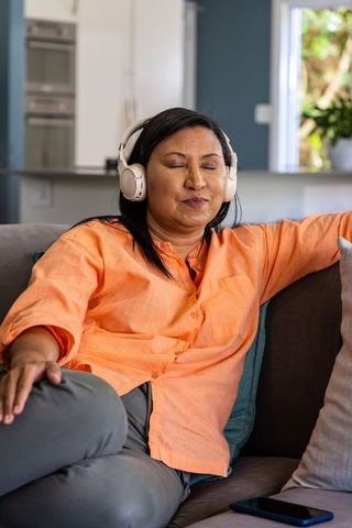 Mature asian woman relaxing on sofa with wireless headphones, enjoying music at home
