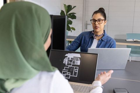 Female coworkers reviewing floor plan on laptop in modern office collaborating on design