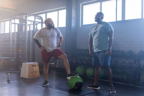 Two african men chatting during training while balancing on medicine ball in gym