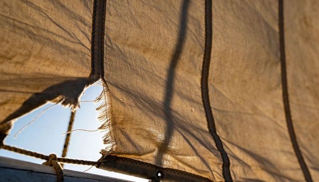Backlit weathered sail with frayed tear and running seams on canvas rigging