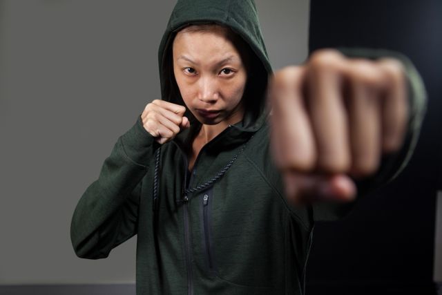 This image portrays a determined woman practicing boxing in a fitness studio. She is throwing a punch while wearing a hooded jacket, exuding strength and focus. The photo is ideal for fitness blogs, gym advertisements, martial arts promotions, and motivational graphics.