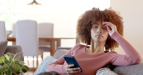 Reclining on sofa holding smartphone in bright living room, cozy home downtime