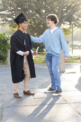 Multicultural graduate celebrating with friend on sunny college campus holding diploma
