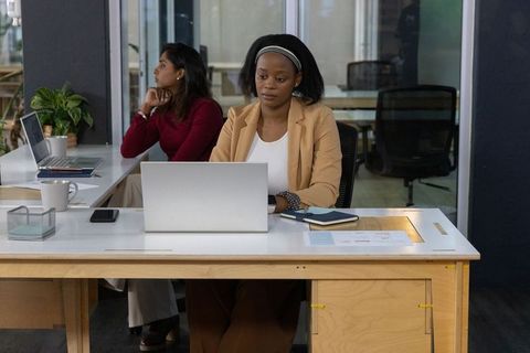 Diverse female coworkers working at modern open office desk using laptops notebooks and coffee