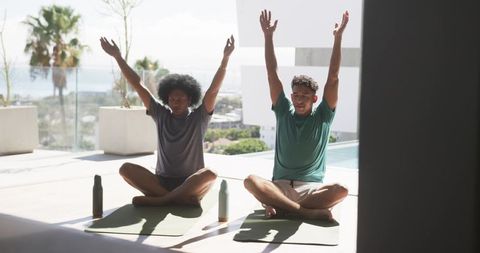 Two men practicing yoga on rooftop terrace raising arms overhead on sunlit poolside mats