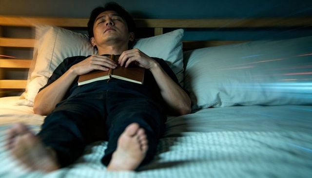 Asian man lying on bed holding book on chest relaxing under soft blue night light
