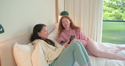 Two friends relaxing on bed in pastel loungewear sharing smartphones and cozy conversation