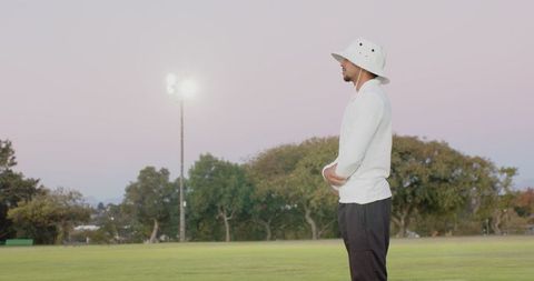 Indian man in bucket hat standing on sports field at dusk smoothing sleeves, thoughtful