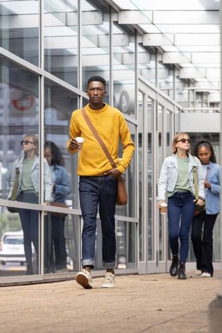 Commuters walking through glass corridor, man wearing mustard sweater holding coffee cup