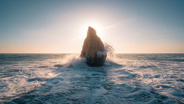 Backlit sea stack rising from ocean at sunrise with crashing waves and spray