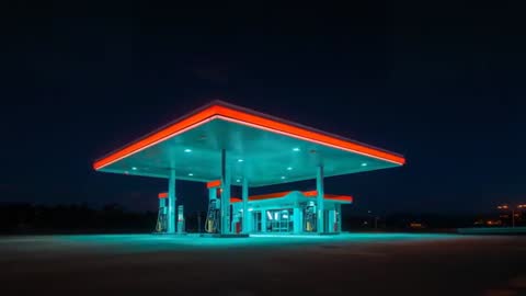 Drone approaching illuminated gas station canopy with teal forecourt glow at night
