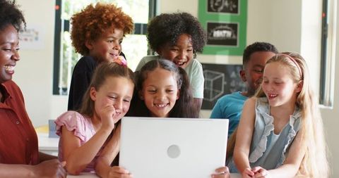 African American teacher guiding diverse elementary students gathering around laptop smiling
