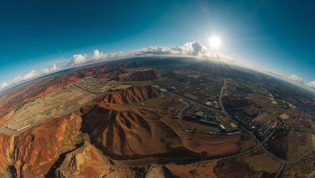 Aerial panorama showing red-rock badlands meeting farmland on arid plateau with sun flare