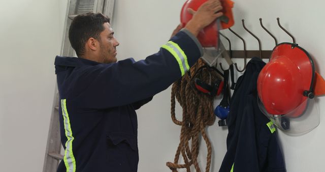Firefighter seen hanging up red helmet and organizing safety gear onto hooks in the fire station. Ideal for use in content related to firefighting services, emergency preparedness, safety routines, and public service promotions.