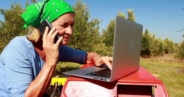 Senior woman in green bandana working on laptop and talking on cellphone in rural outdoor area with fields in background. Ideal for themes of remote work, senior lifestyle, technology use among elderly, and rural occupations.
