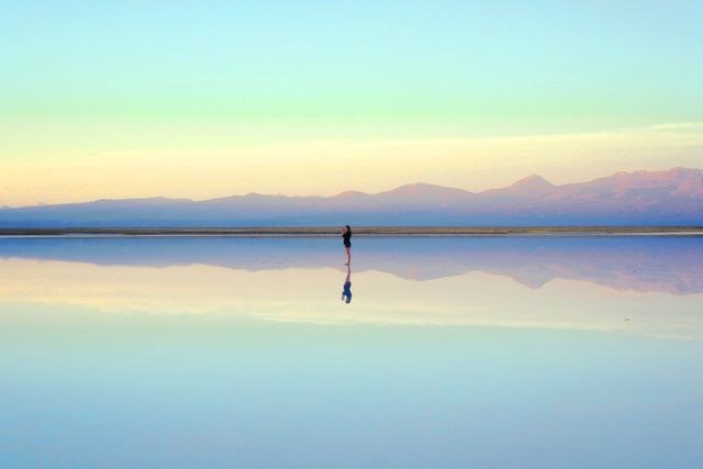 A solitary person stands in the middle of expansive salt flats during dusk. The scene captures the reflection of the individual in the water, creating a mirror effect with pastel colors of the sunset and distant mountain range in the background. Ideal for use in projects related to solitude, reflection, nature, tranquility, and serene landscapes. Suitable as a motivational or inspirational image, desktop wallpaper, or in travel-related content.