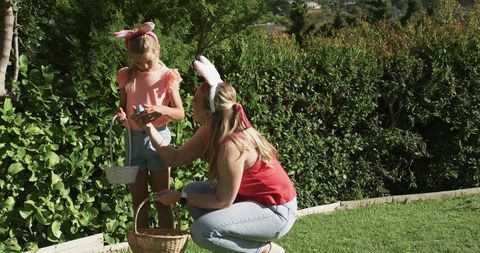 Mother and daughter wearing bunny ears hunting easter eggs in sunny backyard garden