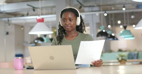 African woman working remotely with laptop and headset holding document in coworking space