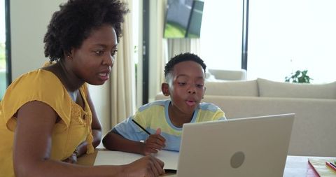 African American mother tutoring son online at home table with laptop and notebook for study