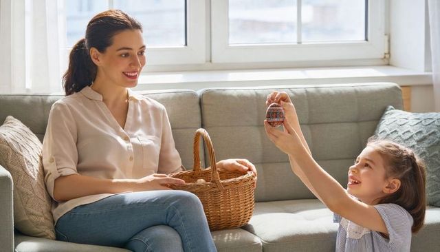 Smiling mother holding wicker basket, daughter showing hand-painted easter egg on sofa
