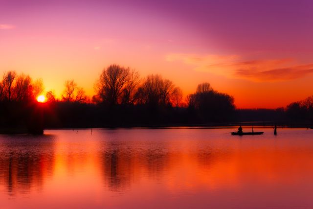 Serene sunset over still lake with bright orange and purple hues reflecting off calm water surface. Silhouette of person in boat adds to calm and tranquil atmosphere. Perfect for promoting relaxation, travel brochures, nature-inspired art, and leisure activities.