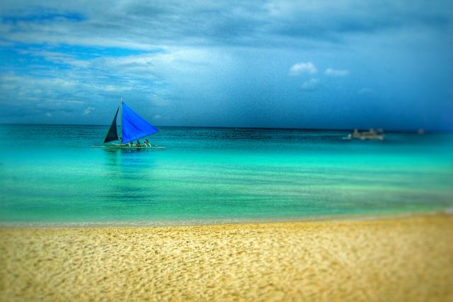 Traditional sailboat gliding on calm tropical waters close to a sandy beach under a cloudy sky. Idyllic seascape featuring vibrant blue and green shades, perfect for use in travel brochures, summer vacation promotions, and stock photography galleries highlighting serene coastal scenes and beach holidays.