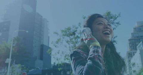 Laughing mid adult Asian woman listening with white headphones on sunny urban street