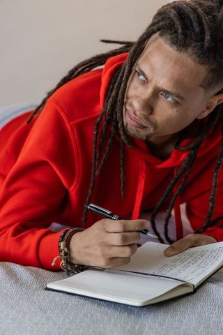 African american man writing in journal while lying on bed in red hoodie