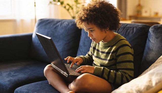 Curly-haired child typing on laptop sitting cross-legged on navy sofa in cozy living room
