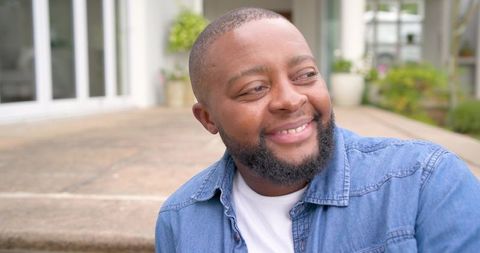 Smiling african man relaxing on stone steps outside home wearing denim shirt and white tee