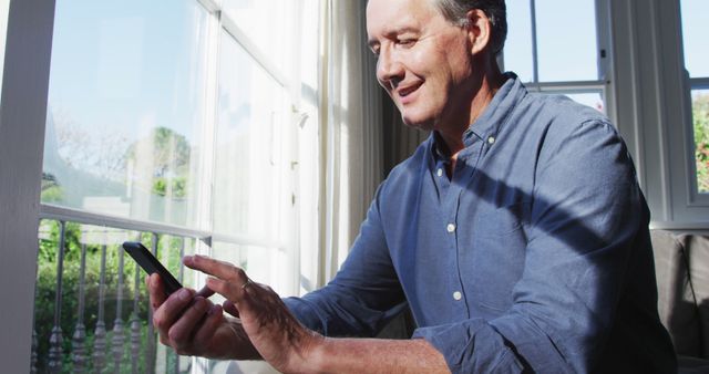 Middle-aged man is using a smartphone near a sunlit window at home. He is relaxed and smiling while interacting with his device. Perfect for concepts like technology, staying connected, casual home settings, or mature individuals embracing digital gadgets.