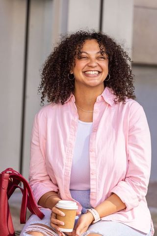 African american woman laughing and holding coffee cup on bench in pink shirt urban casual portrait