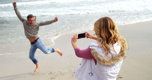 Couple enjoying time at the beach, with one person jumping joyfully while the other captures the moment with a phone. Perfect for travel blogs, vacation advertisements, relationship articles, and lifestyle promotion materials.