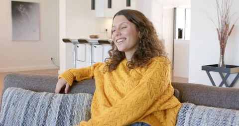 Smiling woman relaxing on sofa in modern apartment wearing mustard yellow sweater