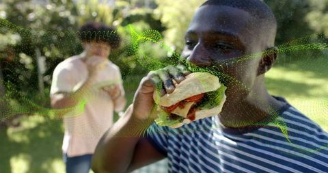 Black man eating sandwich at sunny backyard picnic with green digital particle overlay