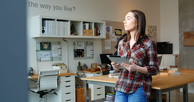 Young adult woman standing in a modern home office, holding a tablet and looking thoughtful. Shelves of books and various office supplies visible in background, suggesting a creative and organized work environment. Useful for illustrating concepts of remote work, creative occupations, modern living, and work-life balance.