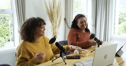 Two female co-hosts recording cozy home podcast using laptop microphones