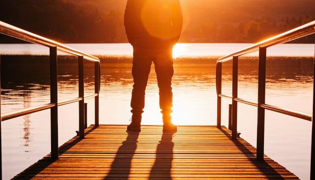Man standing on wooden dock at sunset casting long shadow over calm lake