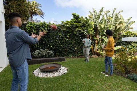 Diverse teenage males tossing american football in sunny backyard with fire bowl, friends enjoying