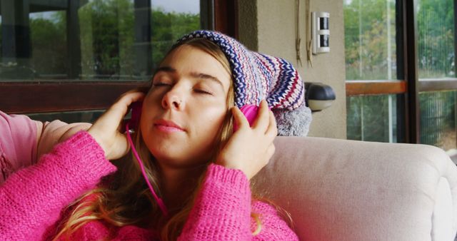 Young woman wearing a knitted hat and pink sweater listening to music with headphones while relaxing on a couch indoors. This image can be used for themes such as relaxation, leisure, comfort, electronic products, lifestyle, or home settings.