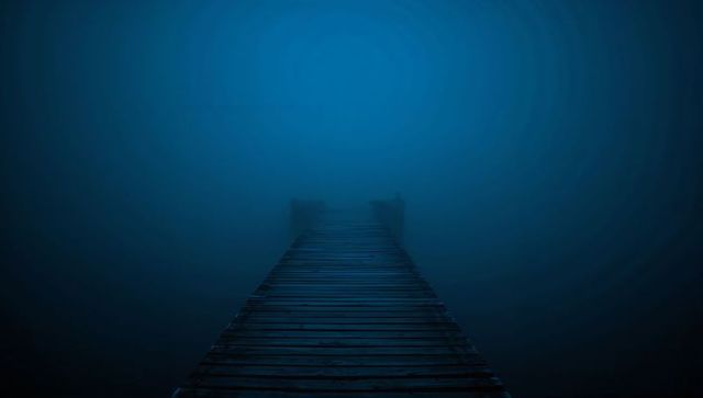 Wooden dock leading into dense blue fog over calm lake, moody minimalist pier at dawn