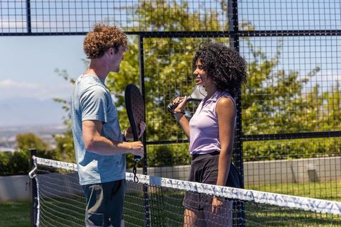Couple chatting at net on outdoor pickleball court holding paddles under sunny skyline