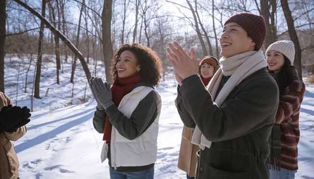Friends clapping and laughing on winter walk in sunlit snowy forest wearing beanies and scarves