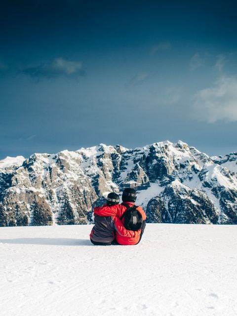 Couple in winter gear sitting on snowy mountain ridge with majestic mountains in background. Perfect for use in travel, adventure, romance promotions, outdoor activity marketing, winter sports advertising, and inspirational nature content.